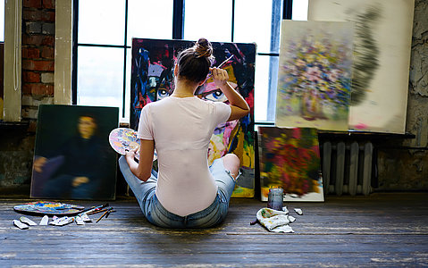 Back view of talented female artist with palette for mixing acrylic colors spending daytime workshop for creating beautiful paintings, inspired woman in casual clothes drawing in loft art studio