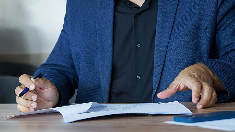 An adult man in a business suit, sitting at a table with a pen, fills out an agreement or order form. Concept of concluding an agreement or filling out forms. Photo. No face. Selective focus