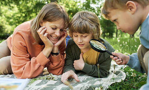 Portrait of mother with two boys using magnifying glass in park and looking at bugs and insects exploring nature together